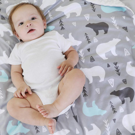 Couverture bébé douce et moelleuse au design nordique, ornée de motifs d’ours blancs, noirs et bleus sur un fond gris, offrant un confort optimal à un bébé souriant allongé dessus en body blanc.