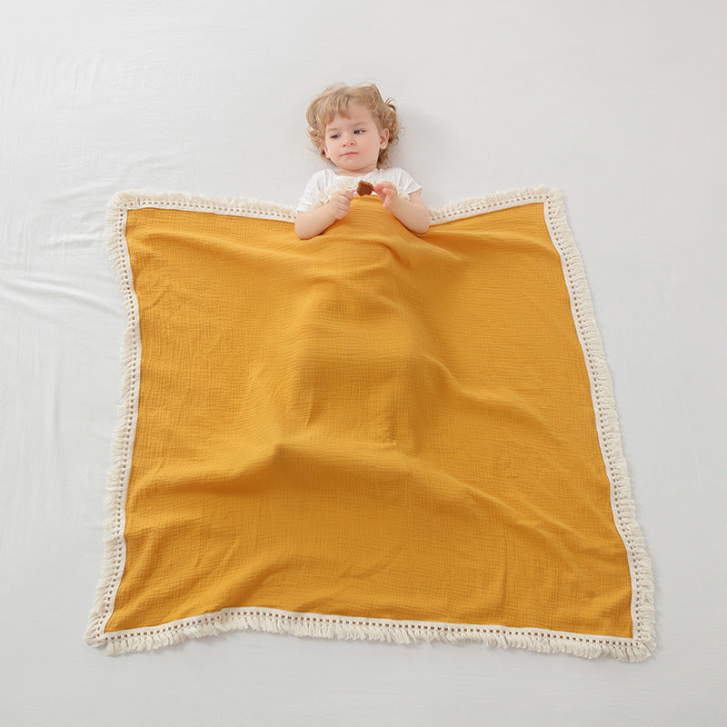 Plaid bébé évolutif en mousseline couleur jaune avec des franges en macramé blanc, recouvrant partiellement un enfant aux cheveux blonds, vêtu d’un t-shirt blanc et tenant un biscuit, allongé sur un drap blanc pour une ambiance douce et minimaliste.