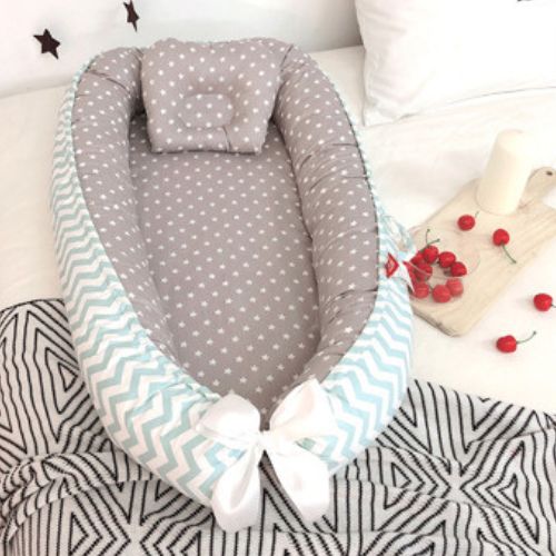 Couffin bébé confortable avec matelas gris à motifs d'étoiles blanches, rebords rembourrés et contour en tissu chevron bleu clair et blanc, agrémenté d'un joli nœud blanc, idéal pour offrir un cocon douillet et sécurisé au nourrisson, posé sur un lit avec une planche en bois décorée de cerises et d'une bougie.