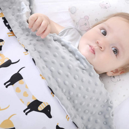 Couverture bébé en tissu ultra-doux avec une face en minky gris à pois en relief et une face imprimée de motifs adorables de chiens, offrant confort et chaleur à un bébé éveillé aux yeux curieux, allongé paisiblement sur un oreiller décoré d’animaux.