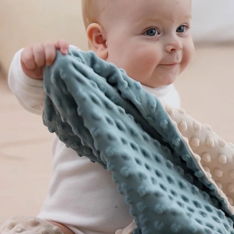 Couverture bébé sensorielle en tissu doux avec motifs en relief, tenue par un adorable bébé aux yeux bleus et au sourire attendrissant, offrant confort et chaleur.