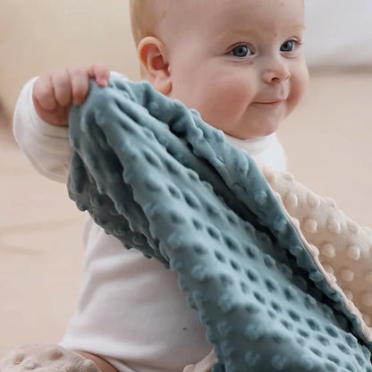 Couverture bébé sensorielle en tissu doux avec motifs en relief, tenue par un adorable bébé aux yeux bleus et au sourire attendrissant, offrant confort et chaleur.