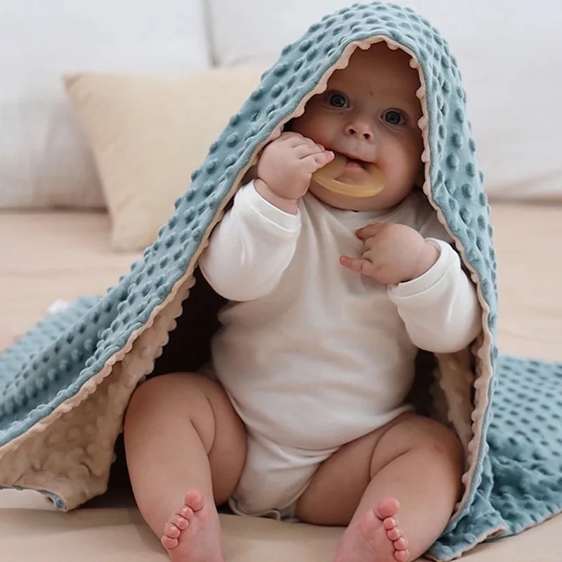 Couverture bébé texturée en tissu gaufré bicolore bleu et beige, fabriquée avec des fibres naturelles et hypoallergéniques, garantissant des matériaux premium pour un confort optimal, délicatement posée sur la tête d’un bébé assis sur un lit, vêtu d’un body blanc, créant une ambiance chaleureuse et réconfortante.