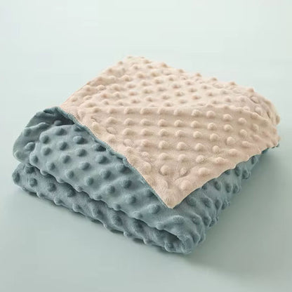 Couverture bébé en tissu doux bicolore, avec une face bleu pastel et une face beige, dotée de motifs en relief en forme de petits pois pour un effet texturé, soigneusement pliée sur un fond uni vert clair.
