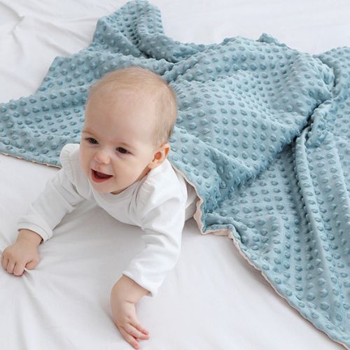 Couverture bébé douce et texturée en tissu gaufré bleu avec revers beige, légèrement froissée et étalée autour d’un bébé souriant vêtu d’un body blanc, allongé sur un lit blanc et s’appuyant sur ses bras en semblant ramper avec joie, créant une ambiance lumineuse et apaisante qui met en valeur le confort et la douceur du textile.