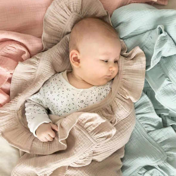 Couverture bébé en mousseline de coton beige avec volants, enveloppant un nourrisson allongé et vêtu d’un body imprimé, entouré de couvertures pastel rose et vert d’eau, offrant douceur, confort et élégance pour le bien-être de votre enfant.