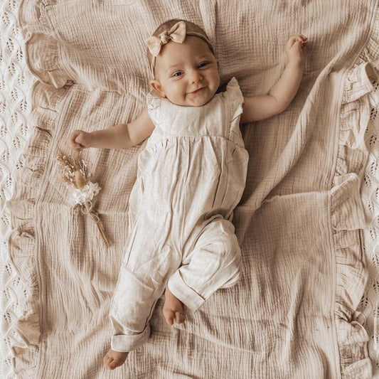 Couverture bébé en gaze de coton beige ultra-douce, parfaite pour envelopper et réchauffer votre nouveau-né, offrant confort et élégance, mise en valeur par un adorable bébé souriant allongé dessus, vêtu d’une combinaison assortie et d’un bandeau avec nœud, accompagné d’un délicat bouquet de fleurs séchées, avec une touche bohème et raffinée.