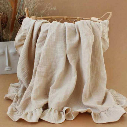 Couverture bébé en mousseline de coton beige avec volants délicats, drapée sur un panier en osier, offrant une texture douce et respirante, idéale pour le confort et la chaleur de votre enfant, dans un décor bohème avec vase artistique et fleurs séchées en arrière-plan.