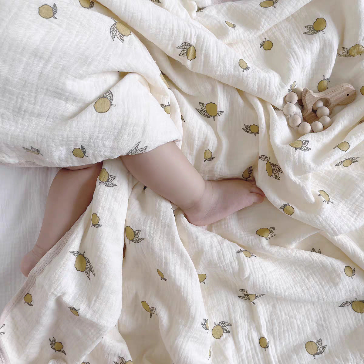 Couverture emmaillotage bébé en mousseline de coton blanche avec un motif de petits citrons jaunes et feuilles vertes, offrant confort et légèreté, recouvrant partiellement un bébé aux jambes croisées et à la peau douce, avec un anneau de dentition en bois naturel et perles en bois posé sur le tissu.