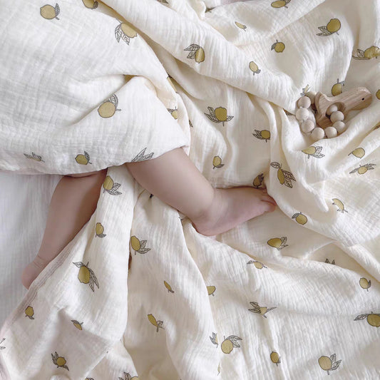 Couverture emmaillotage bébé en mousseline de coton blanche avec un motif de petits citrons jaunes et feuilles vertes, offrant confort et légèreté, recouvrant partiellement un bébé aux jambes croisées et à la peau douce, avec un anneau de dentition en bois naturel et perles en bois posé sur le tissu.