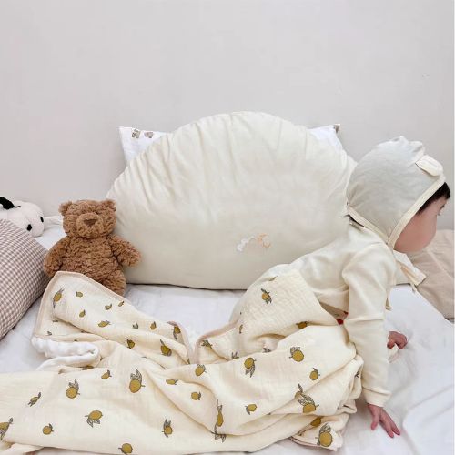 Couverture emmaillotage bébé en mousseline de coton blanche avec un motif de petits citrons jaunes, posée sur un lit aux tons neutres, où un bébé habillé d’un ensemble crème avec un bonnet assorti est en position de ramper. 
