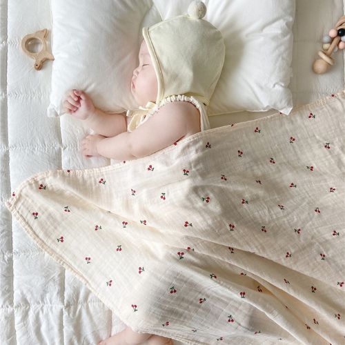 Couverture emmaillotage bébé en tissu beige clair avec un motif de petites cerises rouges et vertes, enveloppant un nourrisson endormi portant un bonnet crème à pompon, allongé sur un lit blanc avec des jouets en bois à proximité.