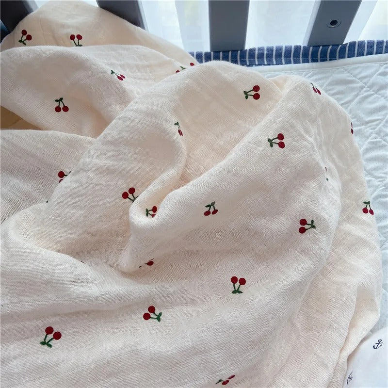 Couverture emmaillotage bébé en mousseline beige, ornée d’un motif brodé de petites cerises rouges avec des tiges vertes, délicatement posée sur un lit près des barreaux d’un berceau.