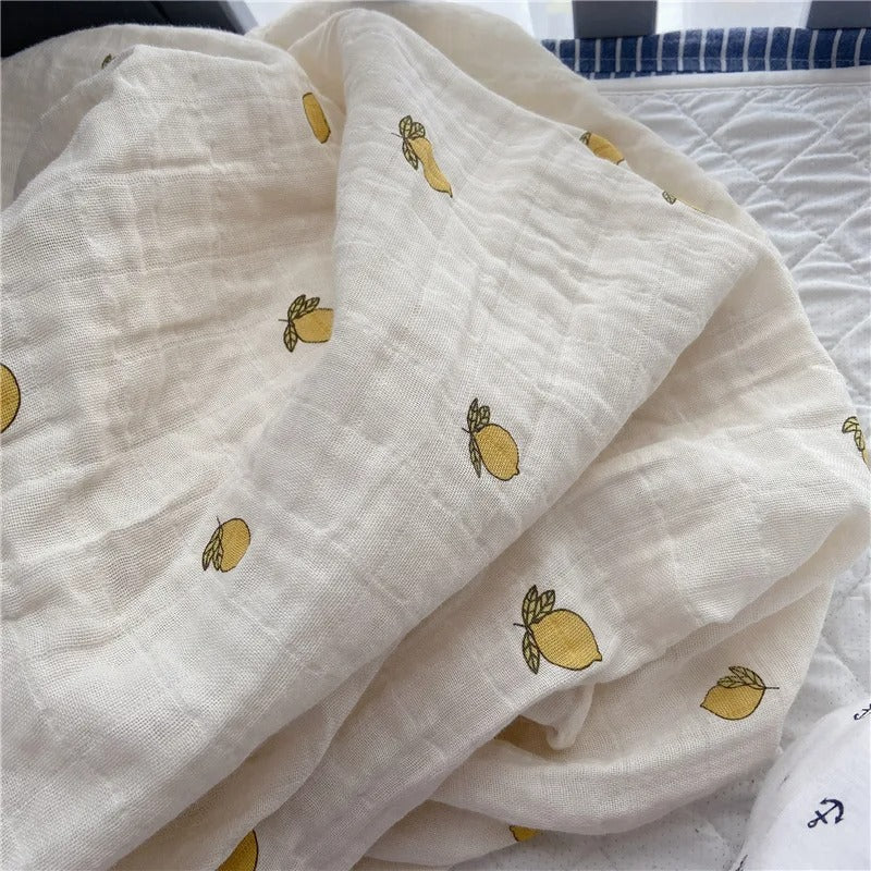 Couverture emmaillotage bébé en mousseline beige, décorée d'un motif brodé de petits citrons jaunes avec des feuilles vertes, délicatement disposée sur un lit près des barreaux d'un berceau.