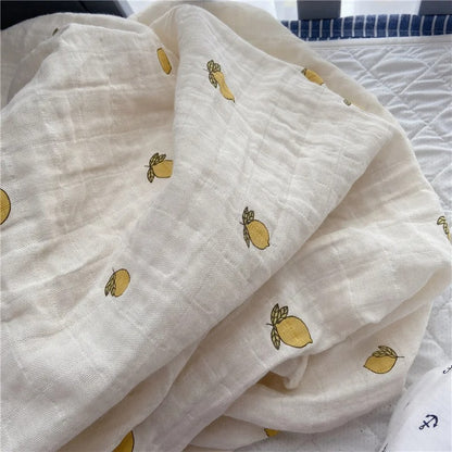 Couverture emmaillotage bébé en mousseline beige, décorée d'un motif brodé de petits citrons jaunes avec des feuilles vertes, délicatement disposée sur un lit près des barreaux d'un berceau.