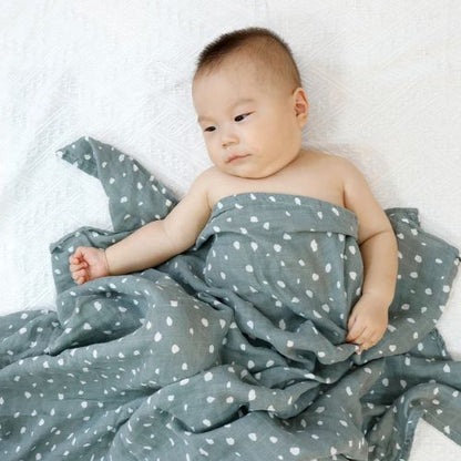 Couverture emmaillotage bébé en mousseline douce de couleur verte avec des motifs blancs en forme de gouttes, enveloppant délicatement un nourrisson allongé sur un drap blanc, offrant confort et douceur tout en apaisant bébé.