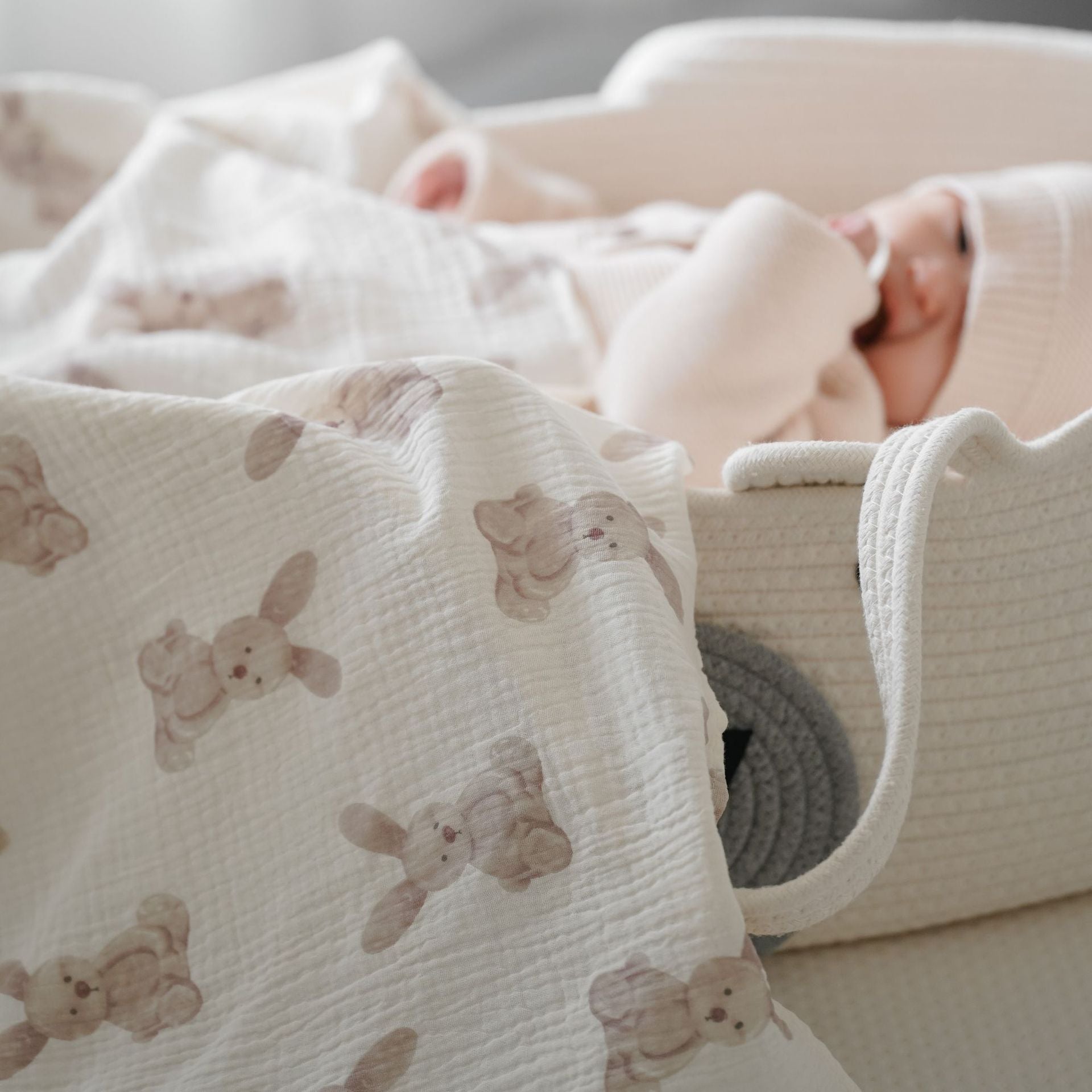 Couverture emmaillotage bébé en mousseline de coton biologique à imprimés lapins beige clair, utilisée dans un couffin tressé avec un nourrisson vêtu d’un ensemble tricoté écru, créant un cocon doux, naturel et rassurant pour les siestes et les moments de repos du nouveau-né.