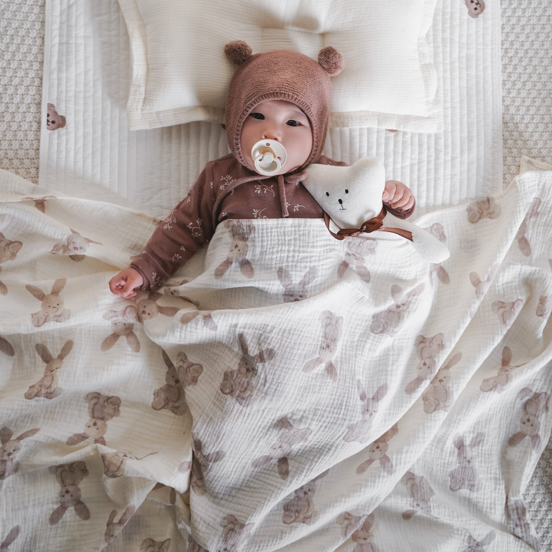 Couverture emmaillotage bébé en mousseline de coton bio ultra douce avec imprimés lapins et oursons beiges, parfaite pour maintenir bébé au chaud durant le sommeil. Bébé allongé sur un matelas moelleux, vêtu d’un bonnet tricoté avec oreilles et d’un pyjama en coton, tenant un doudou en forme d’étoile – une ambiance douce, cocooning et sécurisante idéale pour les siestes et les nuits.