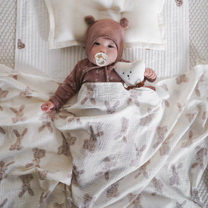 Couverture emmaillotage bébé en mousseline de coton bio ultra douce avec imprimés lapins et oursons beiges, parfaite pour maintenir bébé au chaud durant le sommeil. Bébé allongé sur un matelas moelleux, vêtu d’un bonnet tricoté avec oreilles et d’un pyjama en coton, tenant un doudou en forme d’étoile – une ambiance douce, cocooning et sécurisante idéale pour les siestes et les nuits.