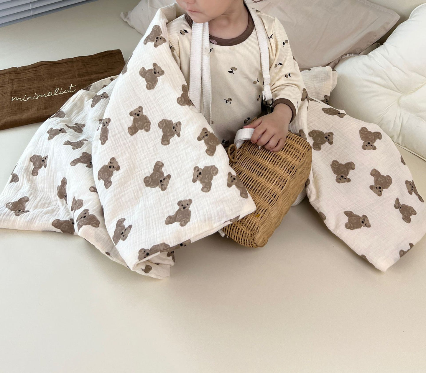 Couverture emmaillotage bébé en coton blanc doux ornée de motifs adorables d’oursons marron, parfaite pour envelopper votre nourrisson avec confort et chaleur, accompagnée d’un bébé en pyjama crème imprimé, tenant un petit panier en osier, dans un univers minimaliste et apaisant.