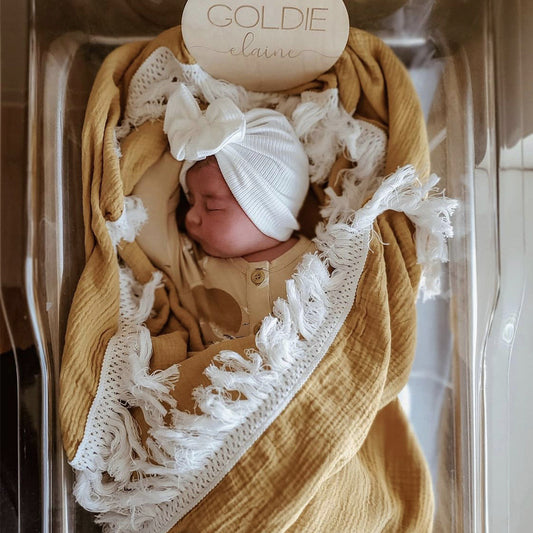 Plaid bébé en coton gaufré couleur Jaune avec franges blanches en macramé, enveloppant un nourrisson endormi vêtu d'un bonnet blanc à nœud et d'une grenouillère beige, allongé dans un berceau transparent avec une pancarte en bois personnalisée portant le prénom "Goldie Elaine".