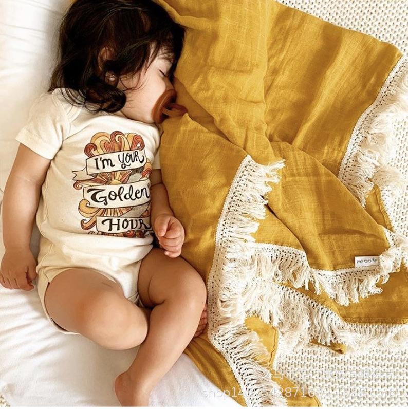 Plaid bébé en mousseline couleur jaune avec franges blanches en macramé, délicatement posé sur un enfant allongé sur un drap blanc avec une tétine ambrée en bouche.