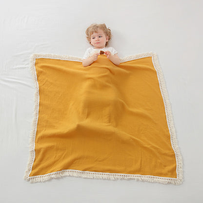 Plaid bébé évolutif en mousseline couleur jaune avec des franges en macramé blanc, recouvrant partiellement un enfant aux cheveux blonds, vêtu d’un t-shirt blanc et tenant un biscuit, allongé sur un drap blanc pour une ambiance douce et minimaliste.