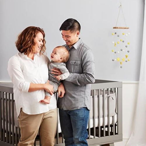 Le tour de lit élégant et sûr, installé sur un lit à barreaux gris, offre protection et style, tandis que de jeunes parents sourient joyeusement avec leur bébé dans un cadre apaisant.