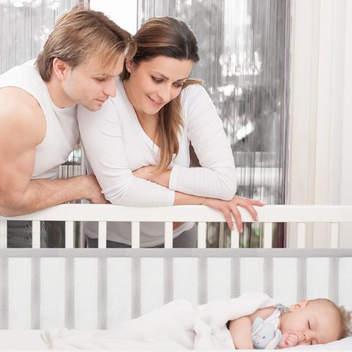 Tour de lit blanc et sécurisé, entourant un bébé endormi paisiblement, sous le regard tendre et souriant de ses parents dans une ambiance lumineuse et sereine.