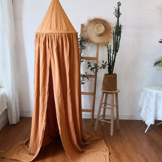 Ciel de lit terracotta en coton suspendu dans une chambre d'enfant avec décoration bohème et tabouret en bois.