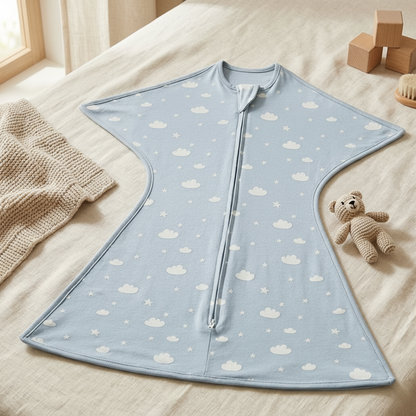 Gigoteuse bébé d'emmaillotage en jersey bleu avec motif nuages et étoiles, posée à plat sur un lit avec un doudou ours et des cubes en bois.