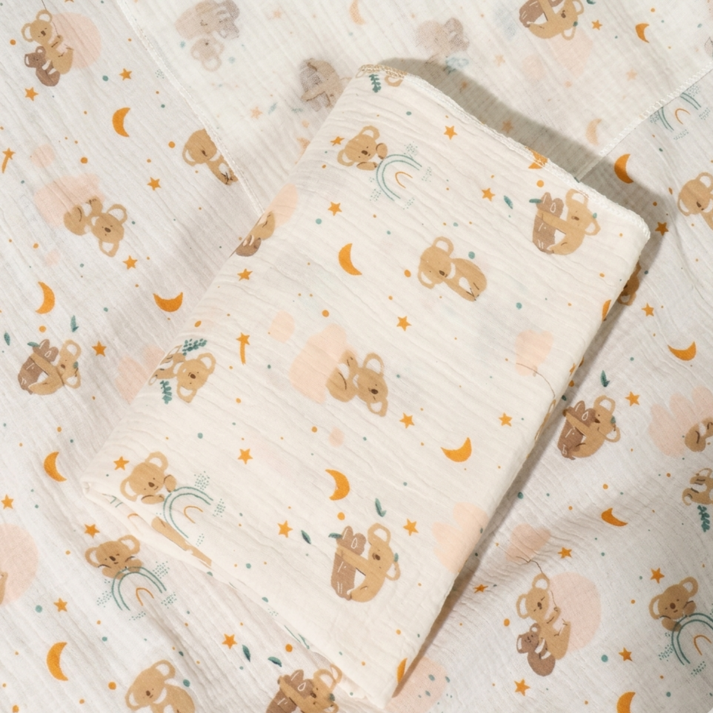 Lange bébé en mousseline de coton blanc cassé avec un motif imprimé de petits koala bruns, de croissants de lune dorés et d'étoiles dorées. Le tissu est doux et froissé, idéal pour l'emmaillotage ou comme couverture légère.
